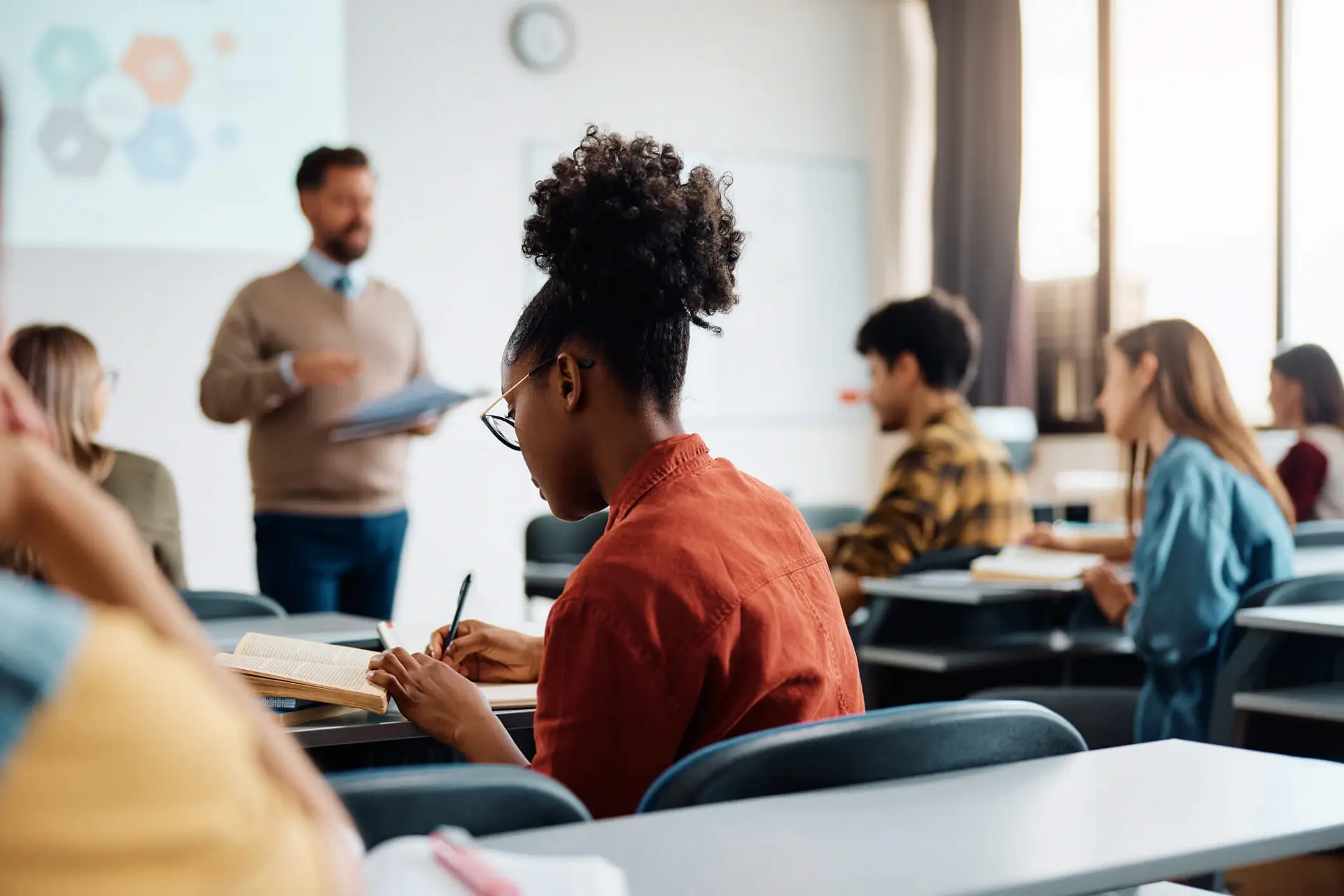 a student sits in a classroom in an orange shirt taking down notes from their teacher