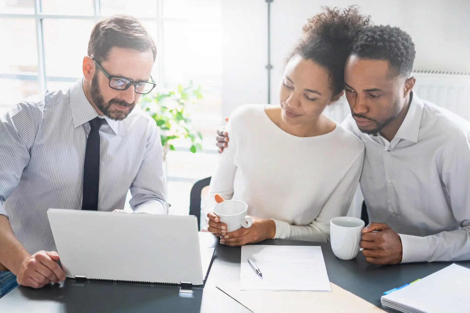 Man on the far left is showing something to a couple on his laptop. The man is wearing a button up white shirt with a black tie. The couple is looking at the screen while cupping their white mugs. The man on the far right has his arm around his partner.