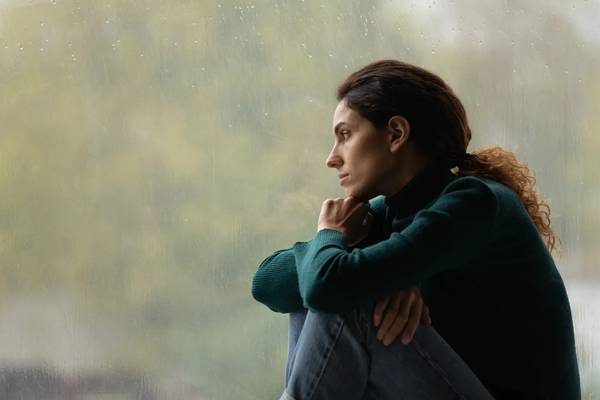 a woman in a green sweater who has possible been sexually abused or assaulted looks out a window contemplating her next steps